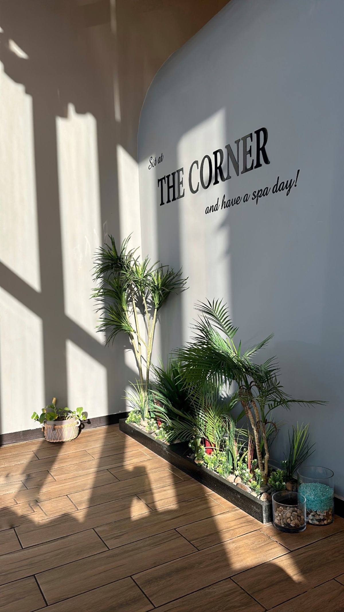 مدخل ذا كورنر سبا: جدار فاتح مكتوب عليه «THE CORNER… and have a spa day!» ونباتات خضراء تحت ضوء الشمس